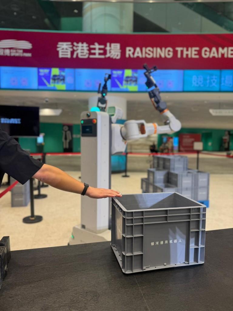 Dual-arm mobile robot at Kai Tak Sports Park with a human hand resting on a crate nearby